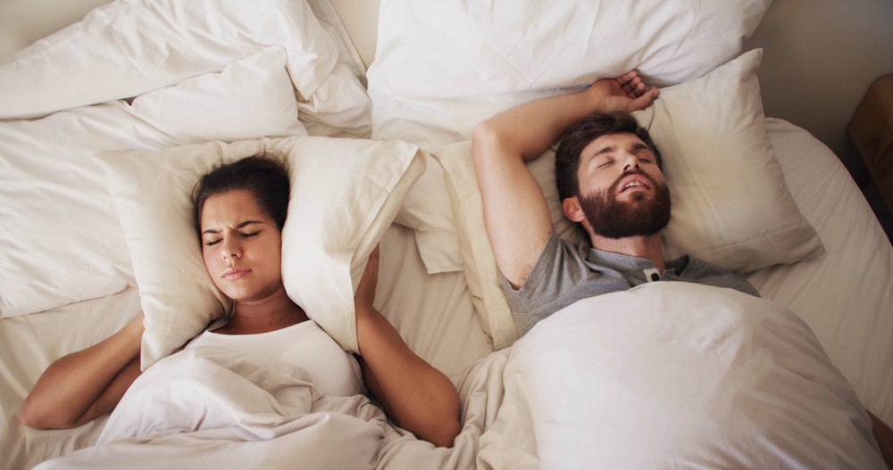 Frustrated woman lying awake in bed unable to sleep while her partner snores loudly beside her, a common experience for couples affected by untreated sleep apnea